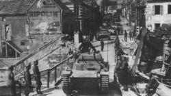 In a captured French town, a German Pzkpfw II grinds across an improvised bridge.